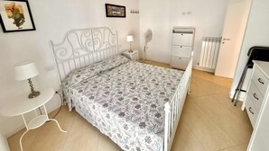 Ein Schlafzimmer mit weißem Metallbettgestell, einem gemusterten Bettbezug, einem Nachttisch mit Lampe, einer weißen Kommode, einem Heizkörper und einer Tür.