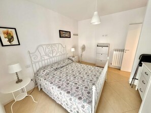 A bedroom with a white metal bed frame, a patterned bedspread, a bedside table with a lamp, a white dresser, a radiator, and a door. - Gianluca apt [center, close to the beach, modern] (Numana)
