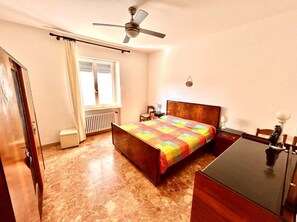 Ein Schlafzimmer mit Bett, Schreibtisch, Stuhl, Deckenventilator, einem Fenster mit Vorhängen und einer Heizung.