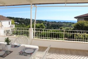 Ein Balkon mit weißer Reling, ein Esstisch und Blick auf eine grüne Landschaft sowie das Meer.