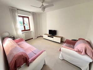 Une chambre avec deux lits, une télévision, un ventilateur de plafond et une fenêtre avec des rideaux.