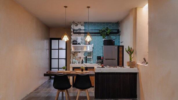 Eine kleine Küche mit Essbereich, ausgestattet mit einem Holztisch, zwei schwarzen Stühlen, Pendelleuchten und einem eingebauten Kühlschrank.