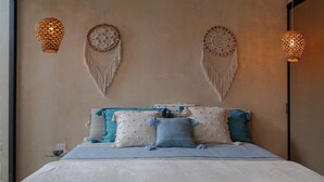 Ein ordentlich bezogenes Bett mit weißer Daunendecke und blauen Akzenten, ein Nachttisch mit Lampe sowie zwei an der Wand befestigte Makramee-Lampen.