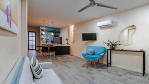Ein modernes Wohnzimmer mit blauem Sessel, weißem Sofa und einem Essbereich, im Hintergrund ist eine Küche erkennbar.