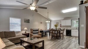 A spacious living room with a sofa, coffee table, and dining area with chairs. A ceiling fan and pendant lights are visible. - A 2-bedroom Hillbilly Getaway house surrounded by charming nature. (Sevierville)