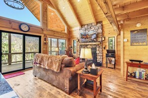 Ein gemütliches Blockhauskaminzimmer mit Kamin aus Stein, Holzbalkendecke, großem Fenster und einer bequemen Sitzecke.
