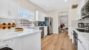 Cocina moderna con armarios blancos, una isla central y un comedor con mesa y sillas de madera.