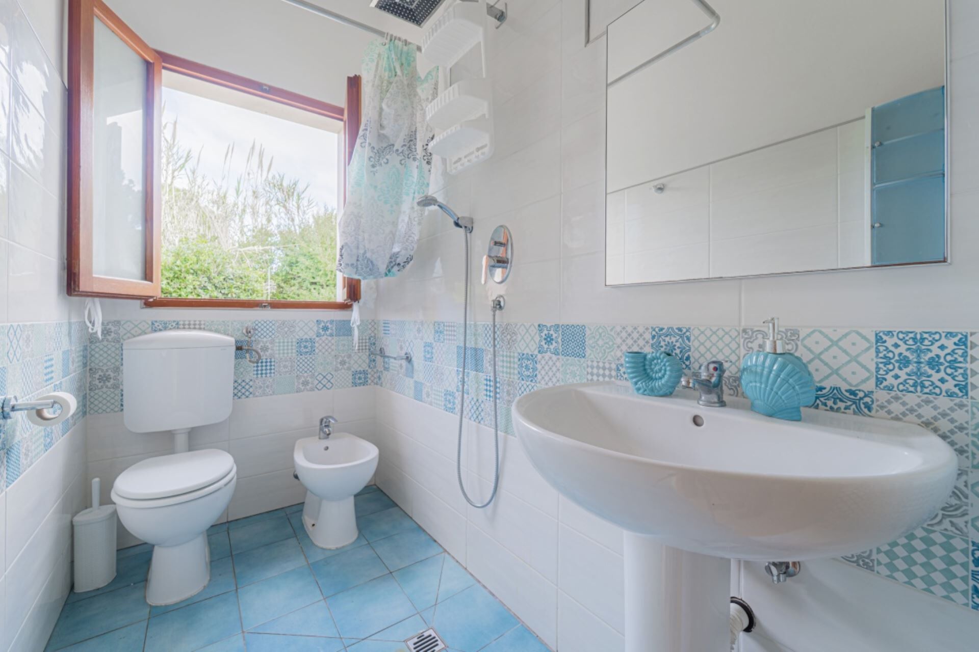 A bathroom with a toilet, bidet, and sink, tiled in blue and white patterns, with a window and a mirror.
