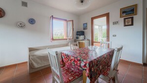 Een eetgedeelte met een tafel, gedekt met een bloemen tafelkleed, witte stoelen, een bank en een raam met gordijnen.