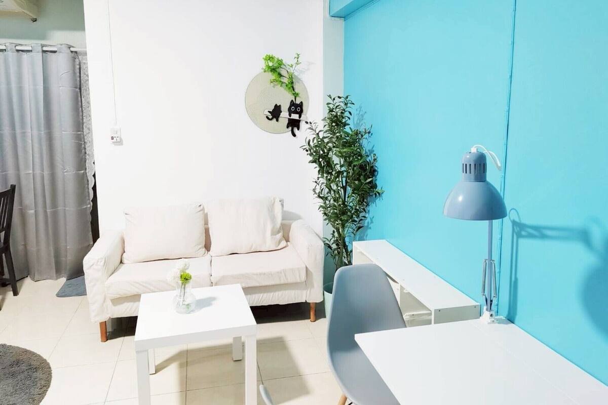 Ein modernes Wohnzimmer mit einem weißen Sofa, einem kleinen weißen Tisch mit einer Vase, einem grauen Schreibtisch und einer grauen Lampe. Eine Zimmerpflanze ist an der Wand, und eine blaue Akzentwand ist sichtbar.