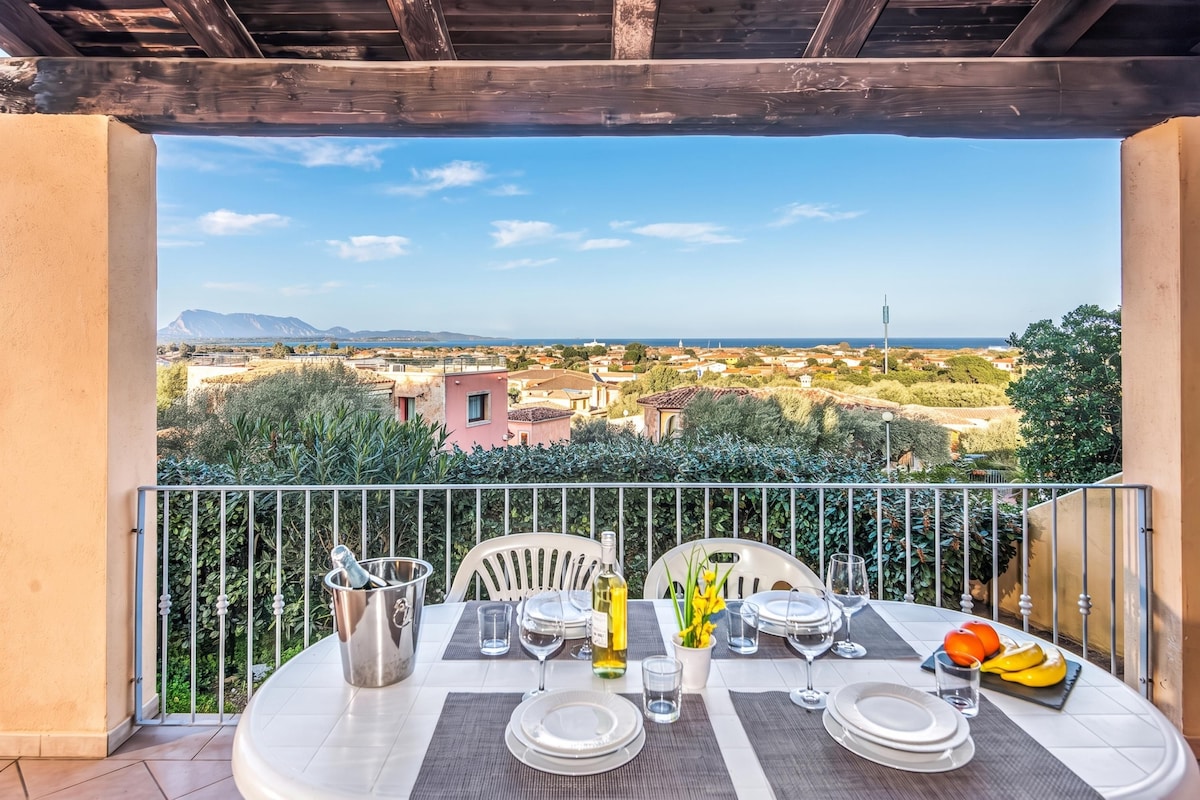 A balcony with a table set for dining, overlooking a coastal town with buildings and mountains in the distance.
