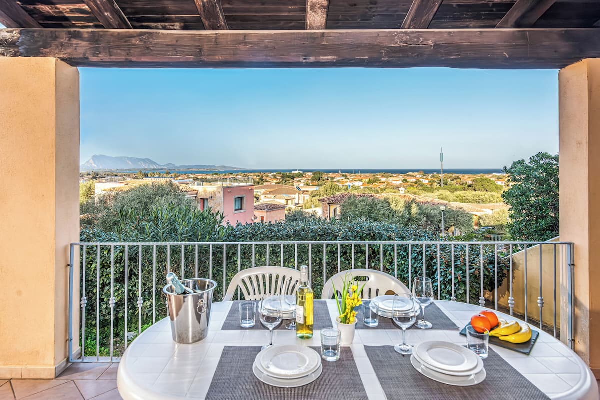 A balcony with a table set for dining, a bottle of wine, and a fruit bowl.
