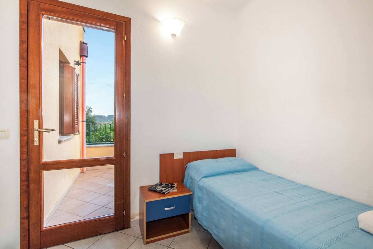 A bedroom with a single bed, a nightstand, a drawer, a wooden door, and a view of the outside.