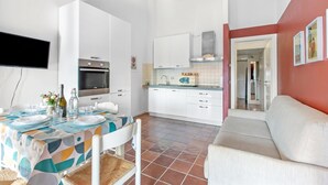 A modern kitchen with white cabinets, a built-in oven, and a microwave. There is a dining area with a table set for two, a sofa, and a TV mounted on the wall. - Cosy House Serena- Park & Shared Pool (Puegnago del Garda)