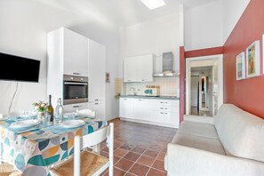 Eine moderne Küche mit weißen Schränken, einem eingebauten Backofen und einer Mikrowelle. Es gibt eine Essecke mit einem Tisch für zwei Personen, ein Sofa und einen an der Wand montierten Fernseher.