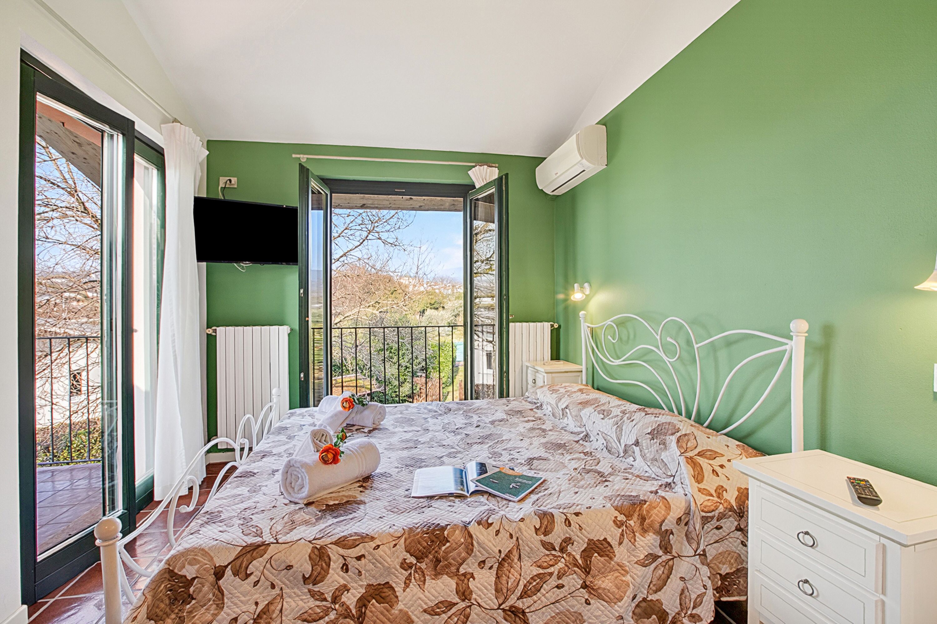 Quarto com cama, mesa de cabeceira, televisão e vista para a varanda.