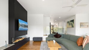 Ein modernes Wohnzimmer mit einem an einer schwarzen Wand befestigten Flachbildfernseher, Holzboden, einem grünen Sofa mit Kissen, einem Couchtisch und einem Kamin mit sichtbarem Feuer.
