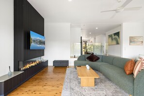 A modern living room with a flat-screen TV mounted on a black wall, a wooden floor, a green sofa with cushions, a coffee table, and a fireplace with a visible fire.