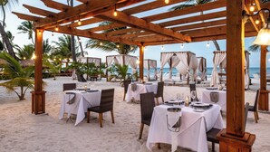 En uteservering med bord för två, placerad under en träpergola som är dekorerad med ljusslingor, med utsikt över en strand med palmer och havet.