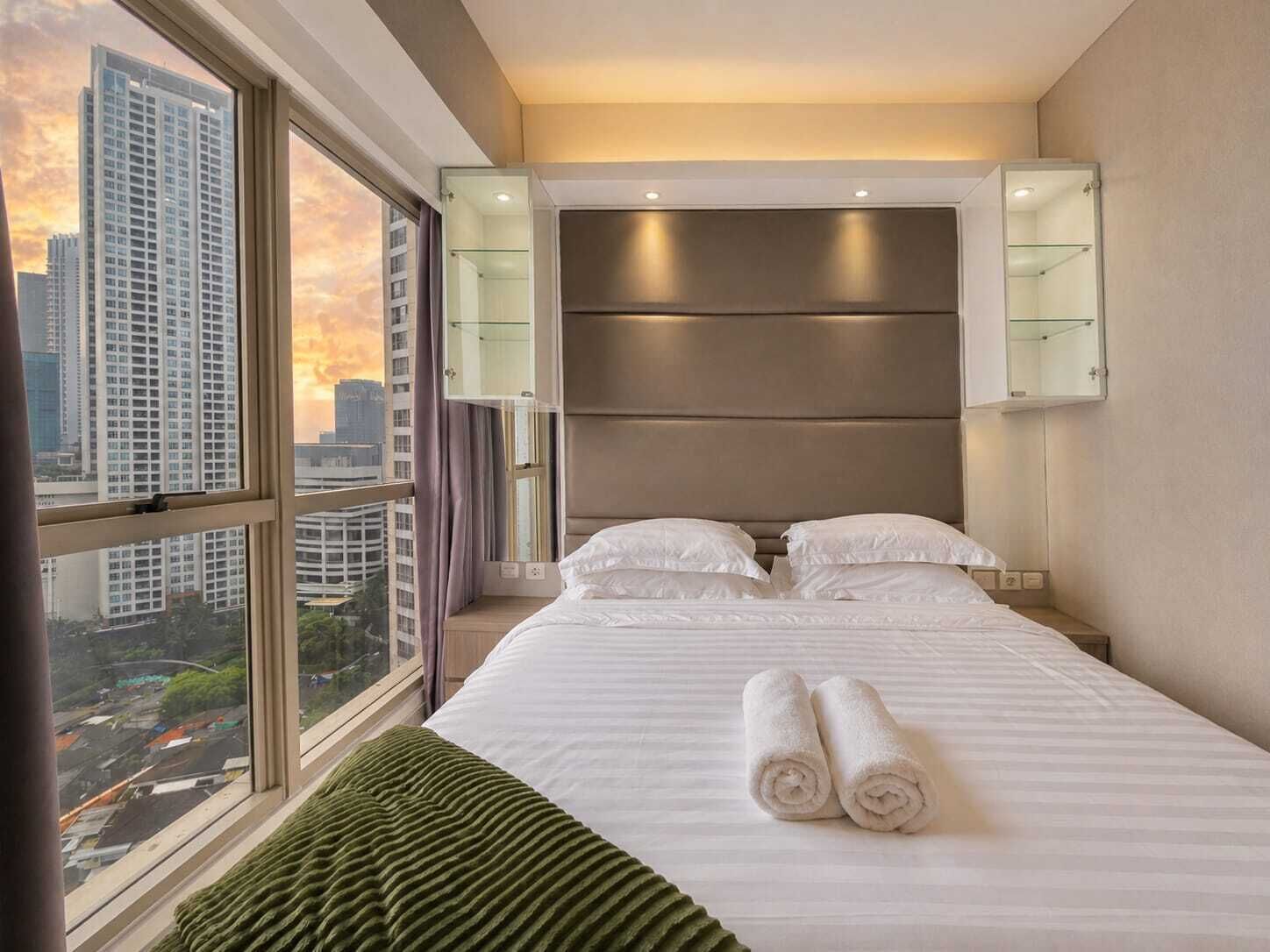 Una camera d'albergo con una grande finestra, un letto con lenzuola bianche, una testiera con illuminazione integrata e vista sulla città.