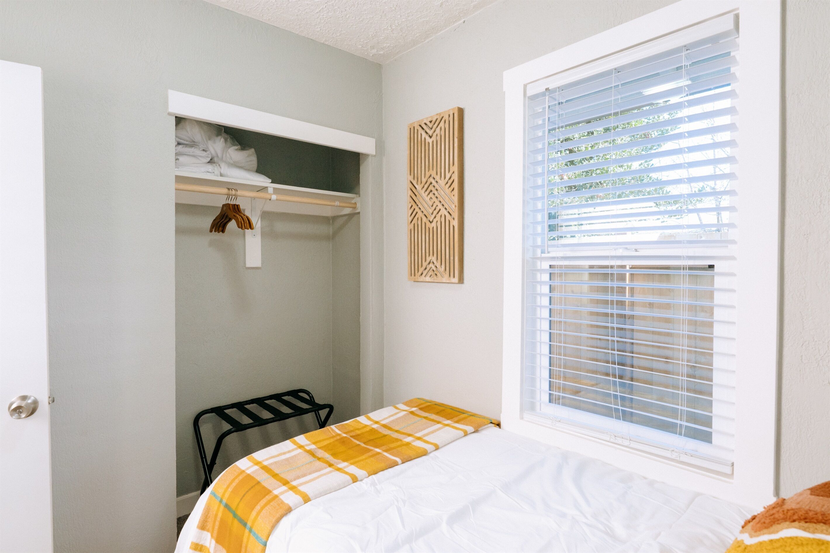 Ein Schlafzimmer mit einem Bett, einem Fenster mit Jalousien, einem Kleiderschrank und einem an der Wand befestigten Regal.