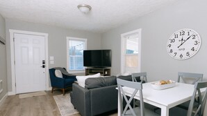 A living room with a grey sofa, a flat-screen TV, a white dining table with chairs, and a wall clock. - Updated 3 Bedroom Charmer (Kelso)