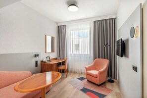 A modern hotel room with a sofa, a wooden table, a desk with a chair, a TV mounted on the wall, and a large window with curtains. - Danubius Hungaria City Centre (Budapest)