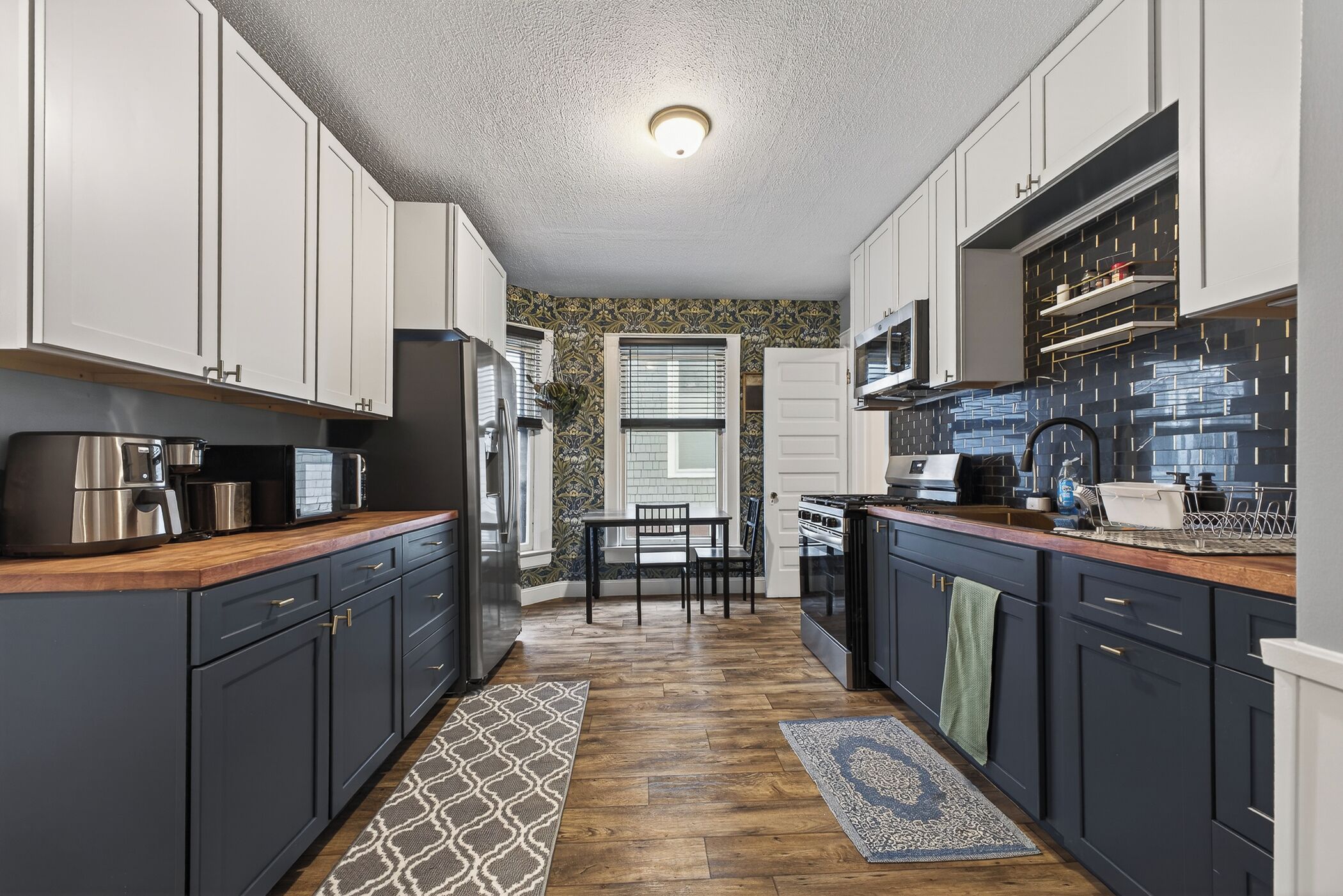 A kitchen with dark cabinets, a wooden countertop, a dining table with chairs, and a patterned rug.