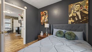 A bedroom with a grey upholstered headboard, a large bed with a grey quilt, two green decorative pillows, a bedside table with a lamp, and a painting on the wall. - Classic Heritage Hill Remodeled 2 Bedroom with Bold and Warm Colors (Grand Rapids)