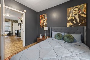 A bedroom with a grey upholstered headboard, a large bed with a grey quilt, two green decorative pillows, a bedside table with a lamp, and a painting on the wall. - Classic Heritage Hill Remodeled 2 Bedroom with Bold and Warm Colors (Grand Rapids)