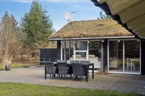A covered outdoor seating area with a table and chairs, a glass door leading to a room, and a satellite dish on a roof. - Lovely holiday home with spa, sauna and peace (Fjerritslev)