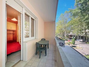 A balcony with a table and chairs, a red bed, and a view of a street with trees and buildings. - Holiday apartment with air conditioning and balcony (Bibione)