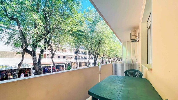 Ein Balkon mit Tisch und Stühlen, mit Blick auf eine Straße mit Geschäften und Bäumen.