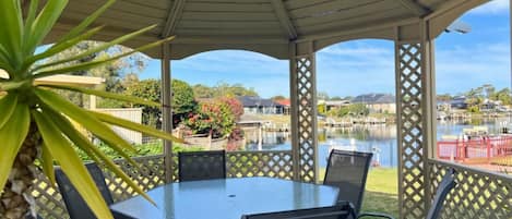 En overdækket terrasse med et bord og stole, med udsigt over vand og boligbebyggelse.