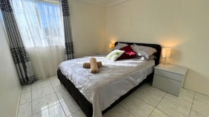 A neatly made bed with a white comforter, two rolled towels, and a variety of pillows. There is a bedside table with a lamp, and the room has a tiled floor. - Chloes Waterfront Oasis ~ A modern apartment with open plan living (Sussex Inlet)