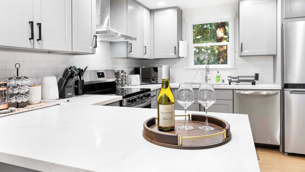 A modern kitchen with white cabinets, stainless steel appliances, and a central island with a wine bottle and glasses. - Newly Modern 4BR2BA Rambler by Super Market (Shoreline)