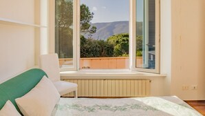 A bedroom with a bed, a chair, and a window with a view of a mountain and greenery. - Villa Giacinto di mare (Capoliveri)