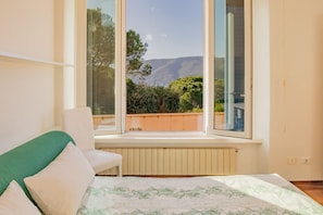 Ein Schlafzimmer mit Bett, Sessel und Fenster mit Blick auf Berg und Grünfläche.