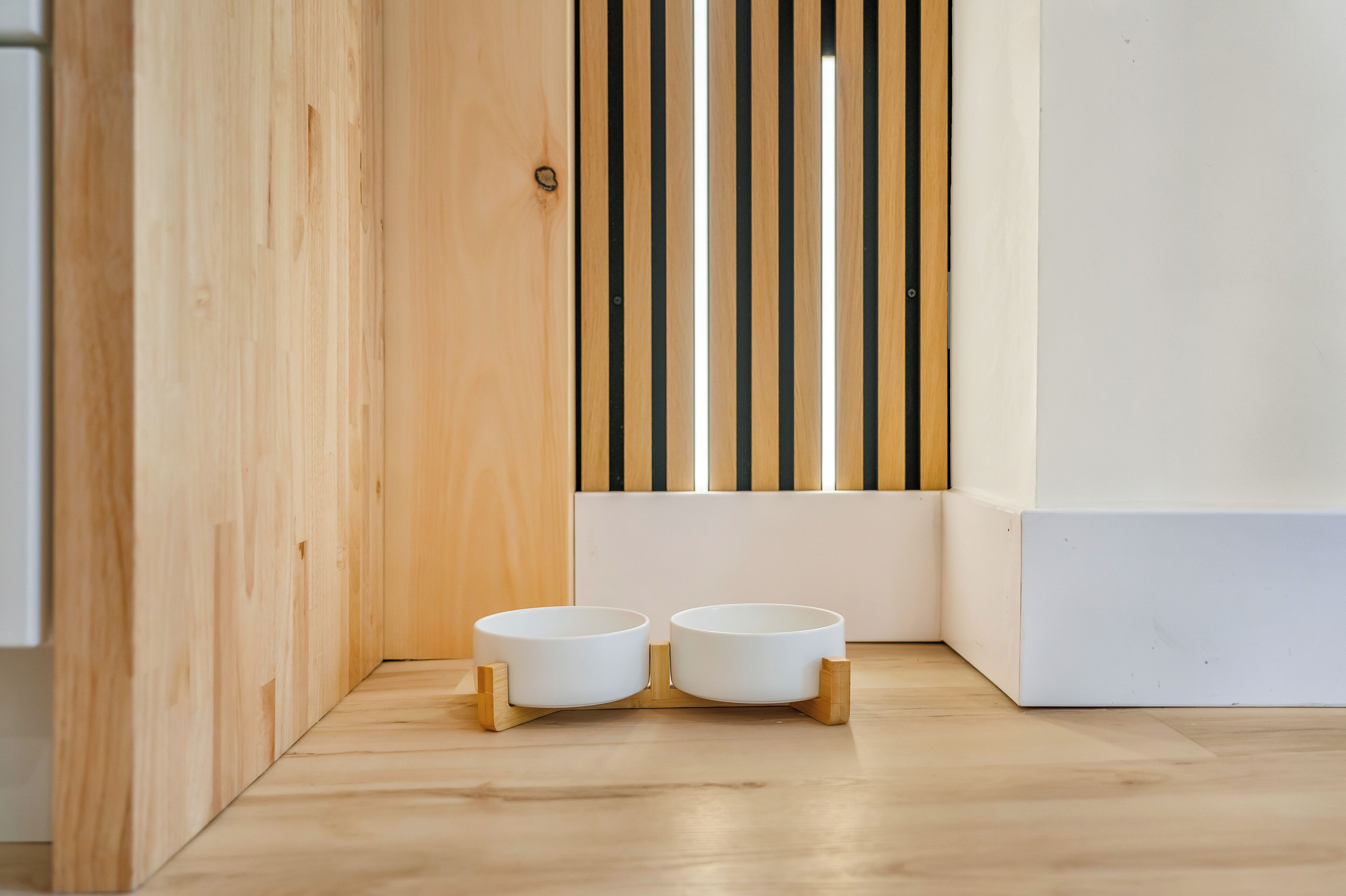 Een hoek van een kamer met twee witte voerbakken op houten standaardjes, een houten lattenwand en een witte muur.
