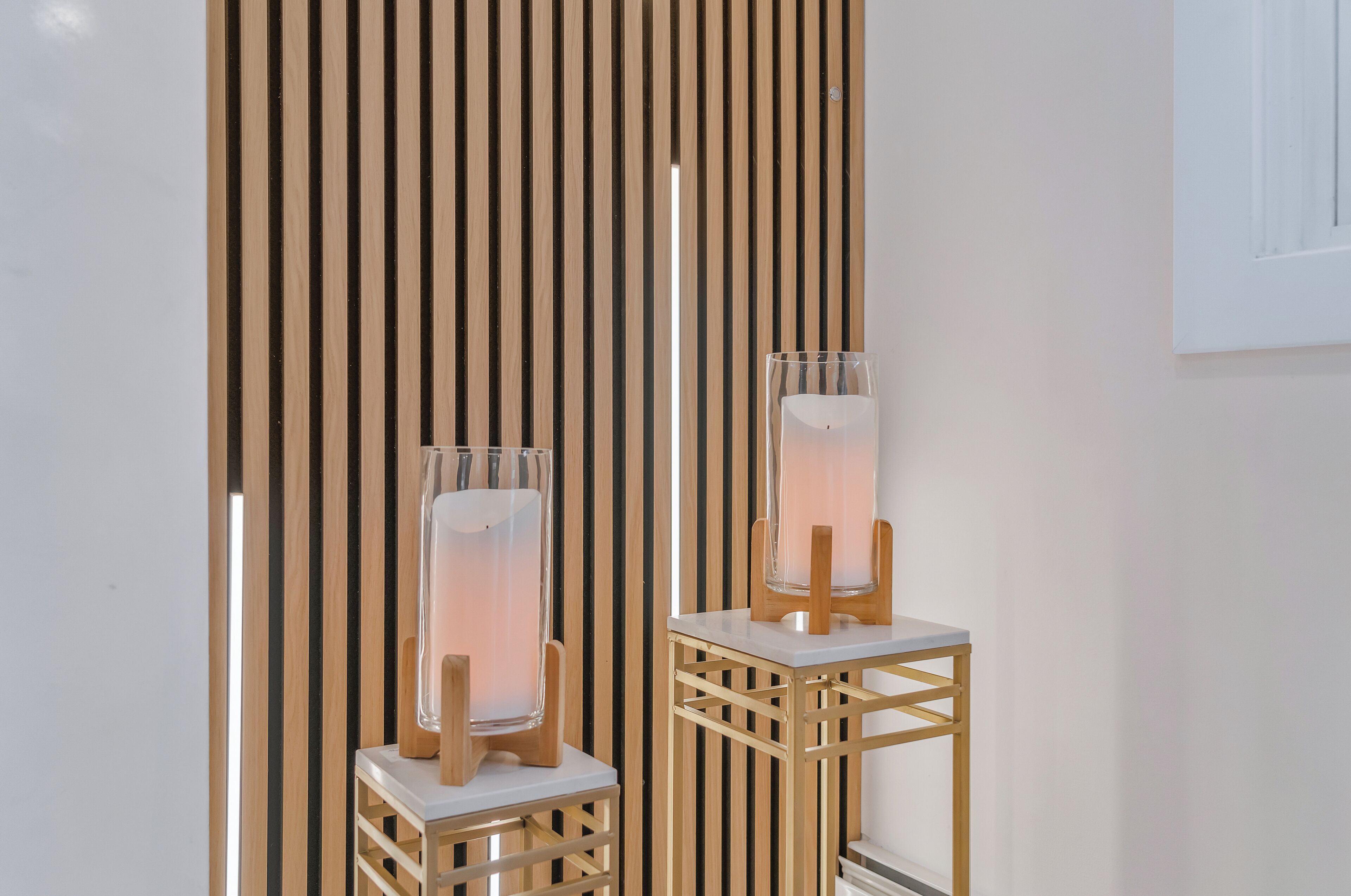 Two glass candle holders on white pedestals against a wooden slat wall.