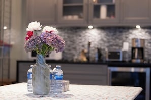 Un jarrón de mármol con flores sobre una mesa, botellas de agua y una cocina al fondo.
