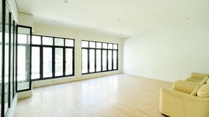 Ein geräumiges Zimmer mit großen Fenstern, einem hellen Holzboden und einem beigefarbenen Sofa.