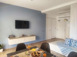 Ein modernes Wohnzimmer mit Fernseher, einem Esstisch mit Frühstücksangebot, einer Couch mit gemusterten Kissen und einem weißen Kleiderschrank.