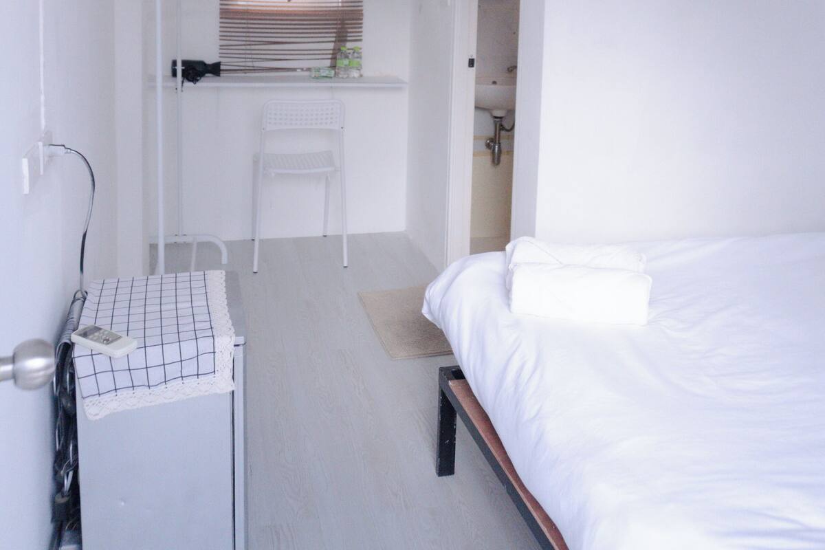 A small, white room with a bed, a desk with a chair, and a window with blinds.