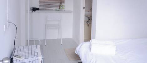 A small, white room with a bed, a desk with a chair, and a window with blinds.