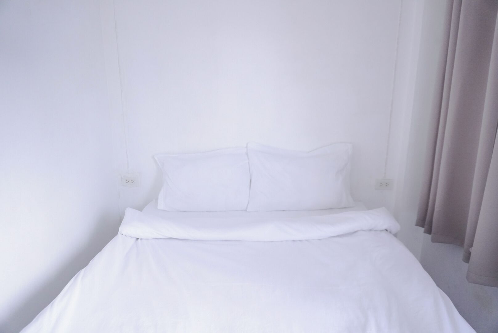Una cama individual con ropa de cama blanca en una habitación con paredes blancas y una ventana con cortinas.