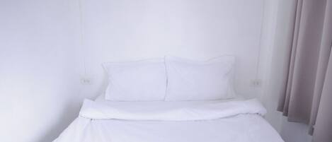 Una cama individual con ropa de cama blanca en una habitación con paredes blancas y una ventana con cortinas.