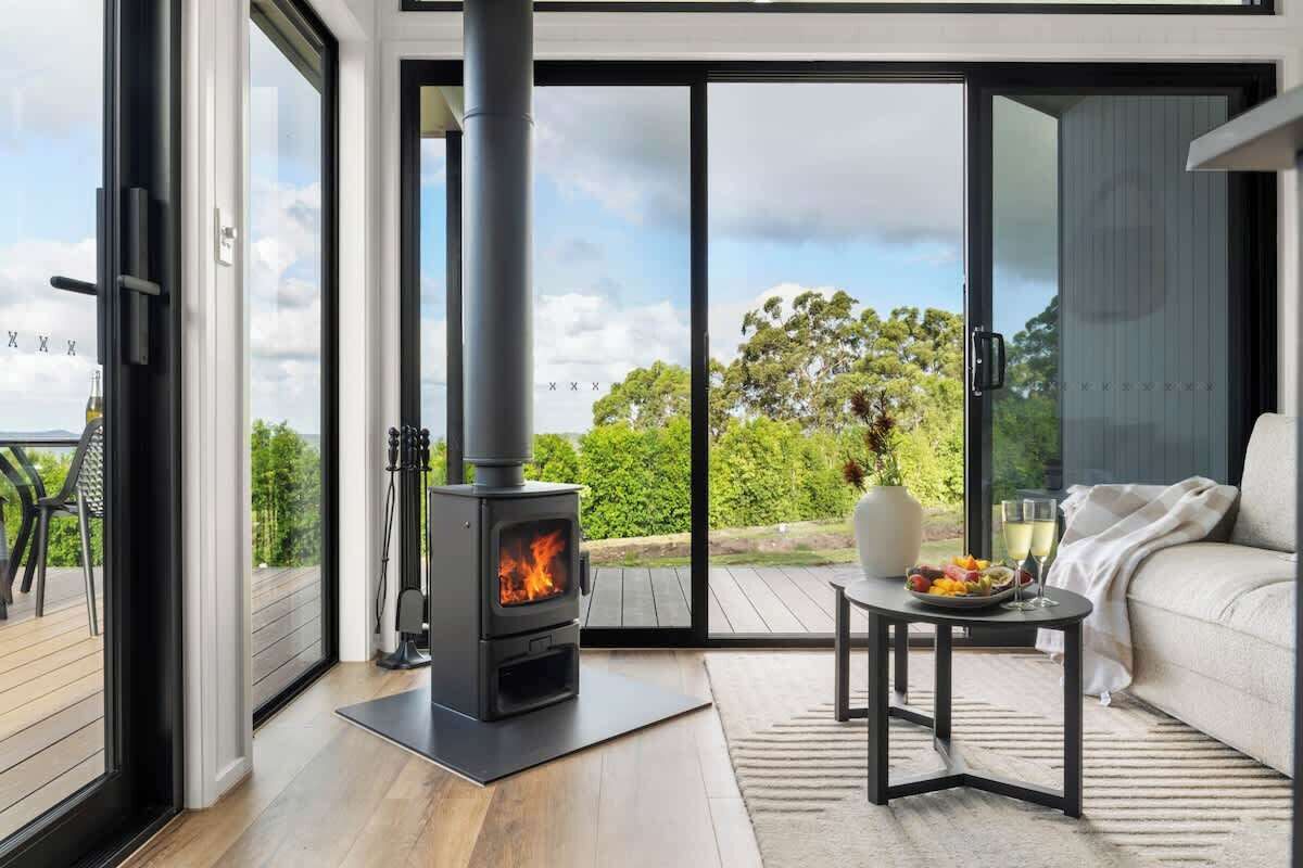 A modern living room with a wood-burning stove, a sofa, a small table with a fruit bowl, and a view of the outdoors.
