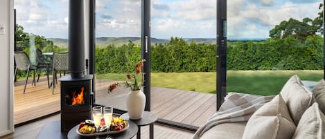 Un salon moderne avec un poêle à bois, un canapé, une table à manger agrémentée de fruits et de boissons, et une vue sur un paysage luxuriant grâce à de grandes portes vitrées.
