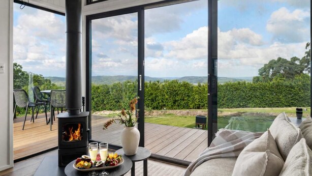 A room with a wood-burning stove, a dining table with a fruit platter and glasses, and a sofa. - Luxury Villa - Firepit - 30min to Most Attractions (Ninderry)